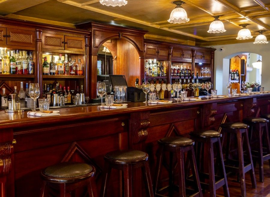 Interior view of a stylish wooden bar featuring an extensive selection of spirits and wines, elegantly arranged glassware, and high bar stools, creating a warm and inviting atmosphere for patrons.
