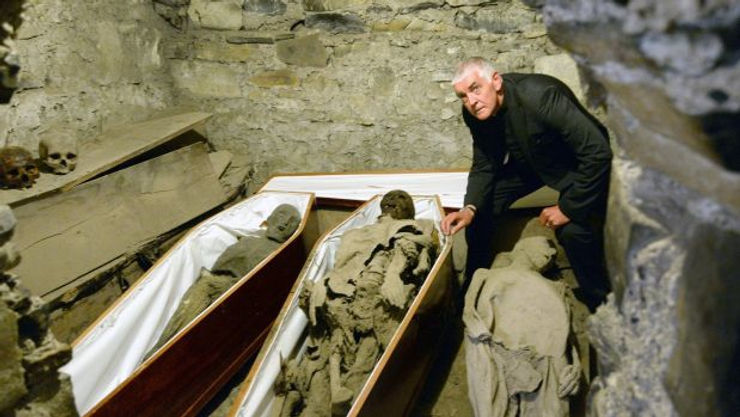 Archaeologist examining ancient mummies in wooden coffins inside a stone crypt, with skulls visible in the background.