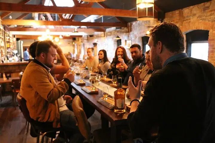 Group of people enjoying a whiskey tasting event at a rustic bar, with a focus on conversation and camaraderie. The setting features wooden beams, stone walls, and various whiskey bottles on display.