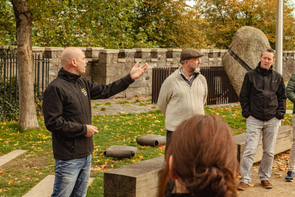 Group of people listening to a speaker during an outdoor tour, surrounded by autumn foliage and historical stone structures. The speaker gestures while discussing local history, engaging the audience in a park setting.