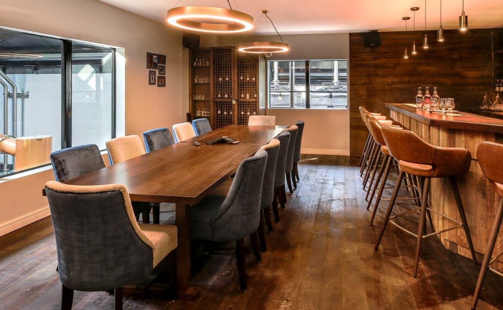 Modern meeting room featuring a long wooden table surrounded by upholstered chairs, with large windows providing natural light and a stylish bar area in the background.