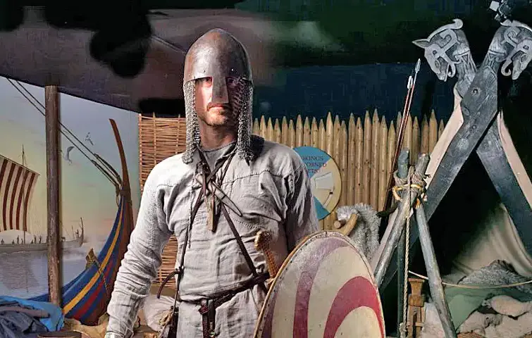 Viking reenactor in traditional armor, including a chainmail coif and helmet, holding a round shield, with a historical Viking camp backdrop featuring longships and wooden fortifications.