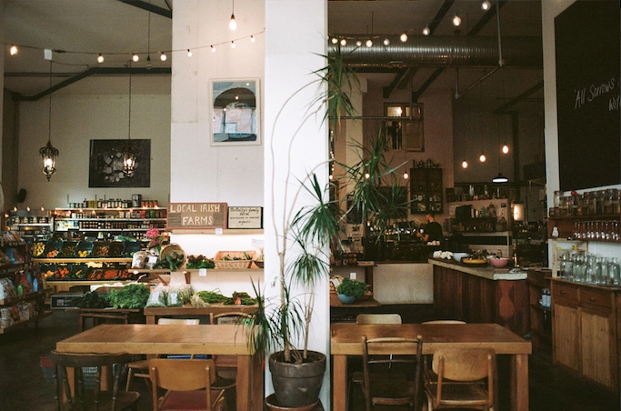 Cozy interior of a local market and café featuring fresh produce displays, wooden tables, and warm lighting. The space includes a sign for "Local Irish Farms" and a coffee bar, creating a welcoming atmosphere for customers.