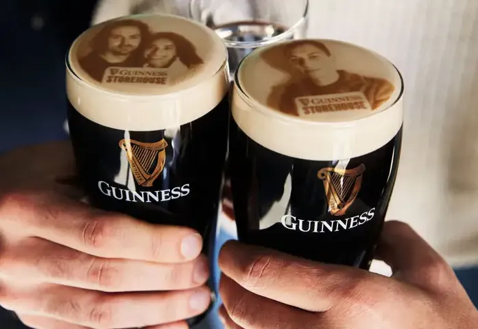 Two hands holding pints of Guinness beer with personalized foam art featuring images on the beer's surface. The glasses prominently display the Guinness logo and are set against a blurred background, creating a cozy atmosphere.