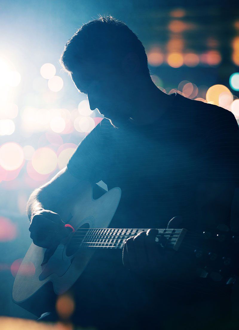 A silhouette of a man playing an acoustic guitar, illuminated by soft, colorful bokeh lights in the background, creating a moody and artistic atmosphere.