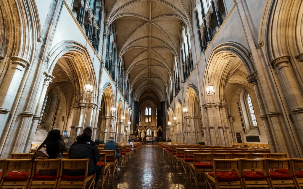Interior view of a cathedral featuring tall arches, elegant lighting, and wooden pews. Visitors are seated, enjoying the serene atmosphere, while stained glass windows add vibrant colors to the space. The architectural design showcases a blend of historical and contemporary elements.