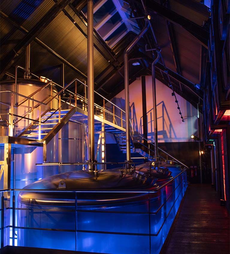Interior of a modern brewery featuring illuminated fermentation tanks, stainless steel equipment, and a staircase. The vibrant blue and orange lighting enhances the industrial atmosphere, showcasing the brewing process in a contemporary setting.