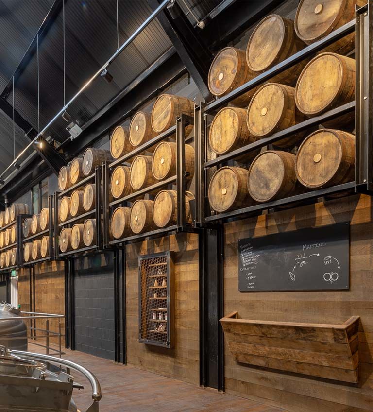 Interior of a distillery showcasing a wall of wooden barrels stacked on metal racks, with a chalkboard detailing brewing notes and a rustic wooden shelf nearby. The industrial design highlights the craftsmanship of barrel storage in a modern setting.