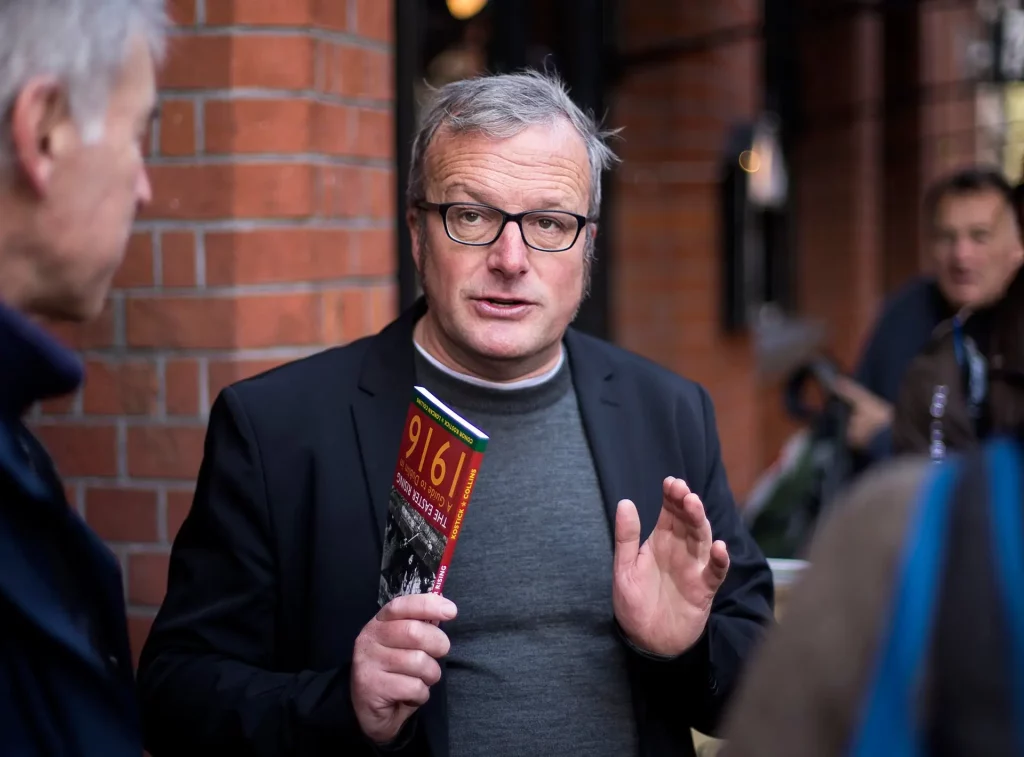 A man with glasses wearing a black blazer and gray sweater gestures while holding a brochure. He appears to be engaged in conversation outside a brick building, with people in the background.
