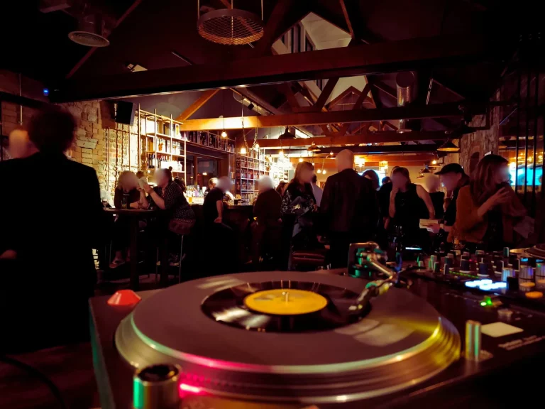 A vibrant nightlife scene featuring a turntable in the foreground, with patrons enjoying drinks and socializing at a bar in a stylish venue illuminated by warm lighting.
